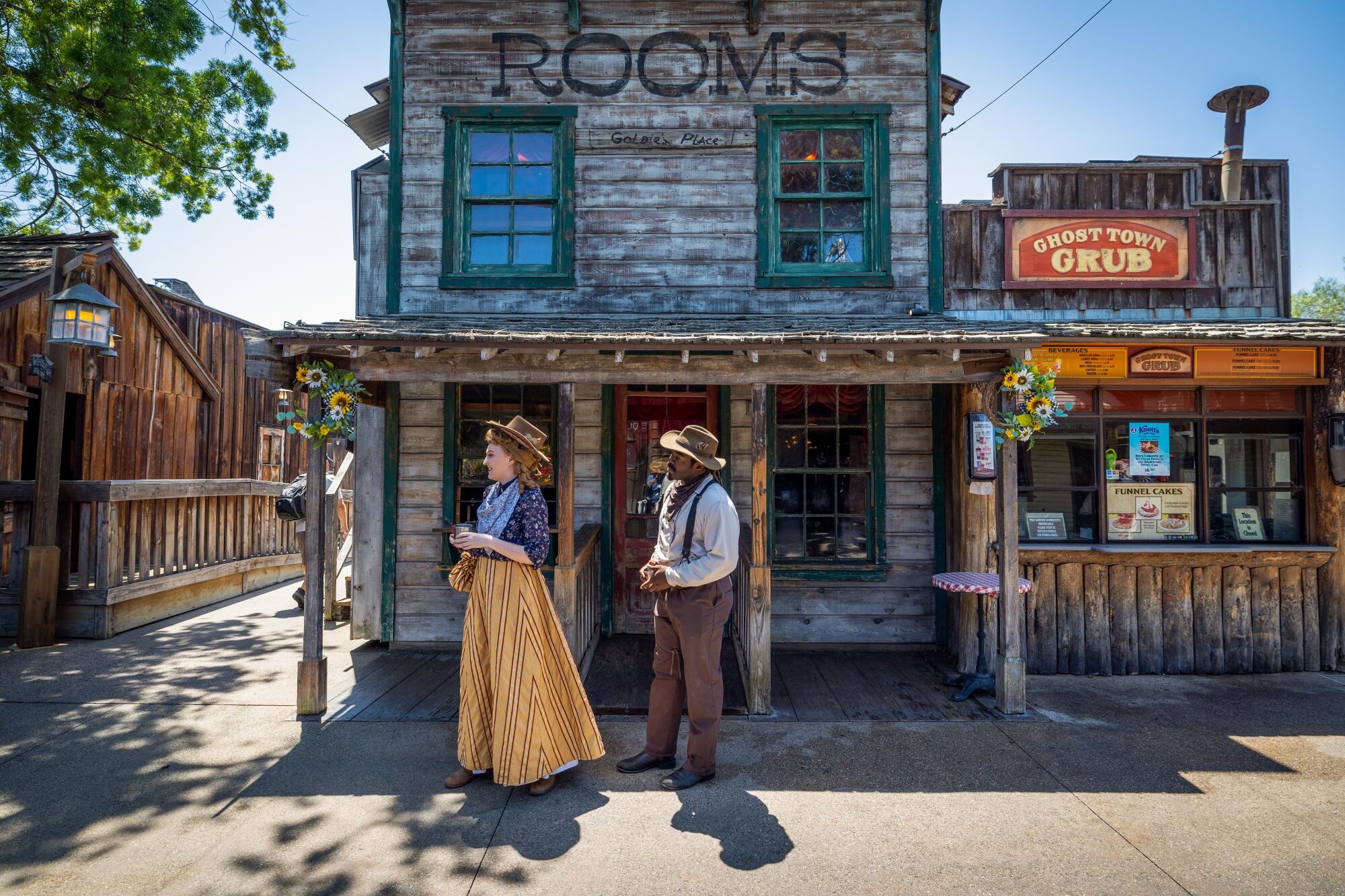 Two people in western-style period clothing in front of an old-looking wooden building