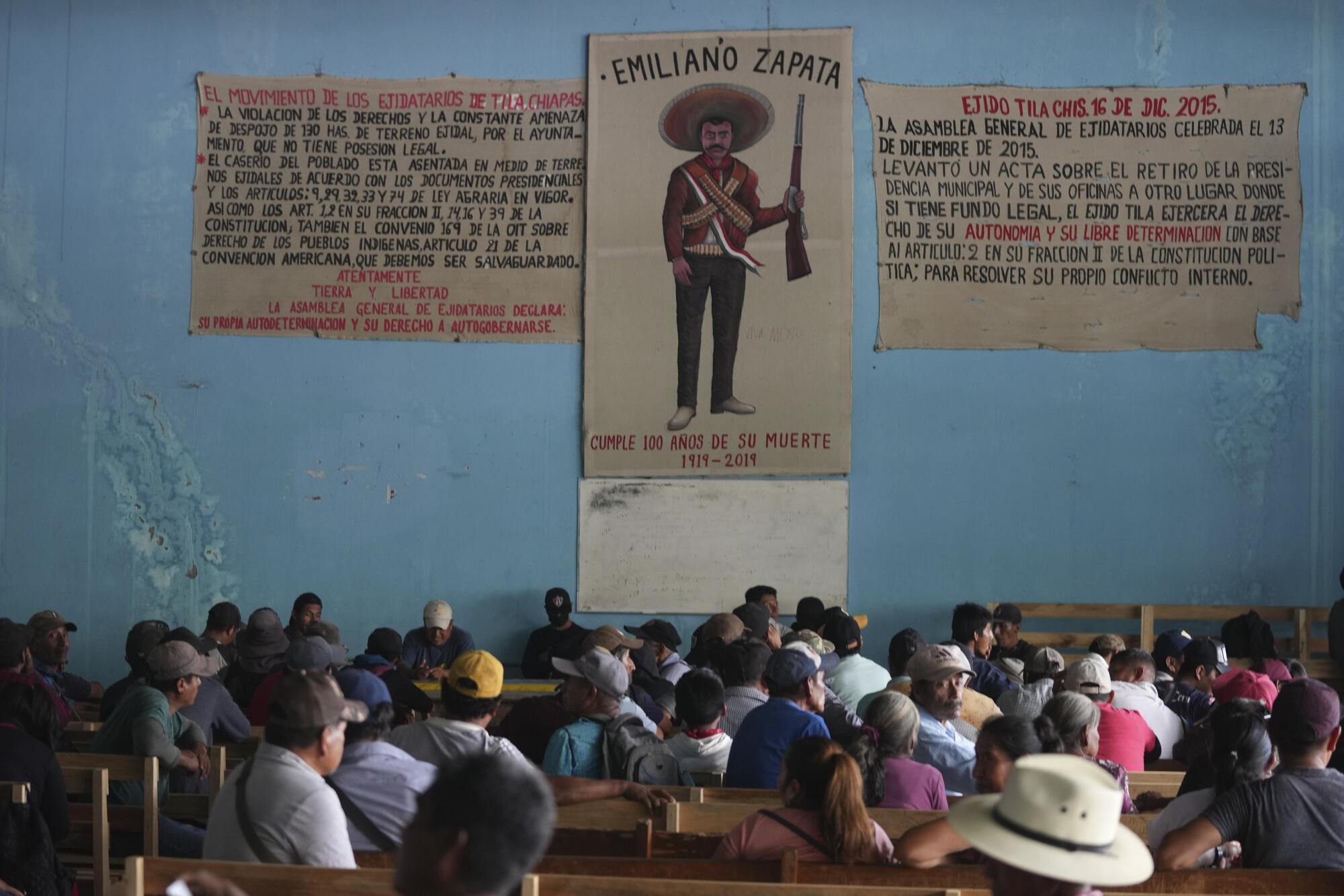 People seated in a room with blue walls and a poster of a man in a hat with a gun and the words Emiliano Zapata